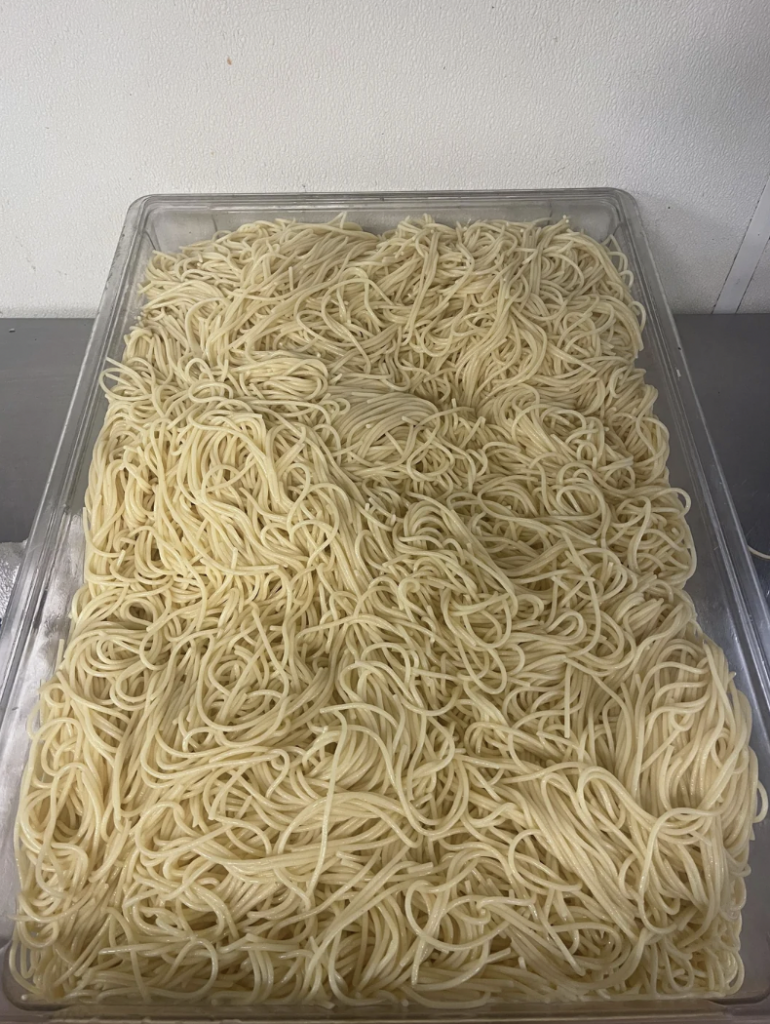 A large, transparent container filled with cooked spaghetti noodles is placed on a countertop. The noodles are light yellow and arranged in an irregular heap, with some strands loosely overlapping others. The background is a plain white wall.