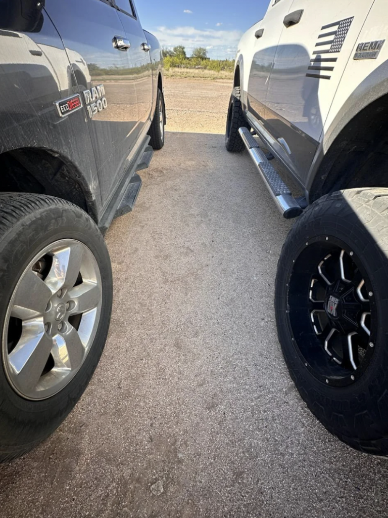 A close-up shot of two parked pickup trucks side by side on a dirt surface. The truck on the left is a gray RAM 1500, and the truck on the right is a white vehicle with unique black wheel designs and decals, including an American flag. The tires are prominently visible.