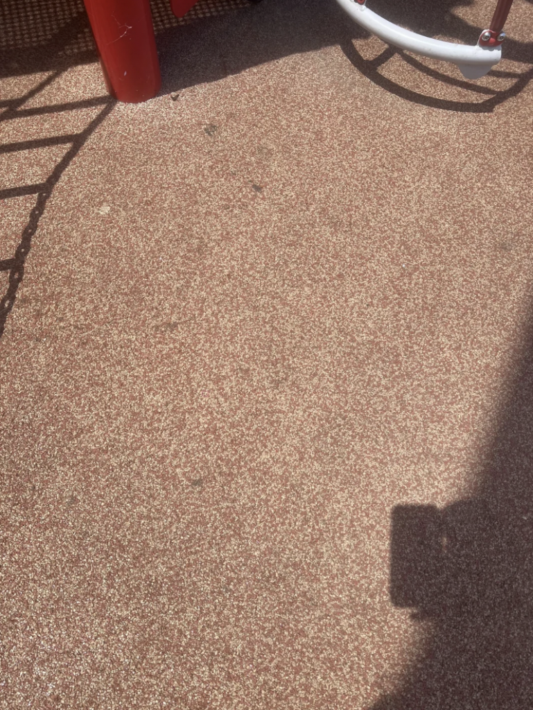 A sunlit patch of red and white speckled ground surface, partially shadowed on the right. The corner of a white and red playground structure is visible on the upper left. The shadow of a person holding a phone or camera is on the bottom right corner.