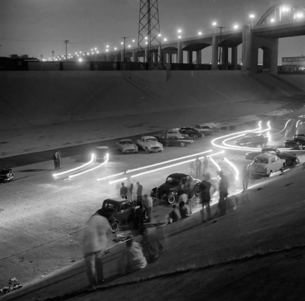 A black-and-white photograph depicts a nighttime scene with several vintage cars parked beneath a bridge. People are gathered around the cars, and long-exposure light trails from moving vehicles create bright, curved lines across the image.