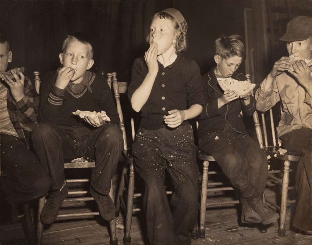 A group of five children sit on wooden chairs eating popcorn. The children, wearing casual clothes, appear engrossed in their snacks, with some having spilled popcorn on their laps and the floor. The scene seems to capture a candid, nostalgic moment.