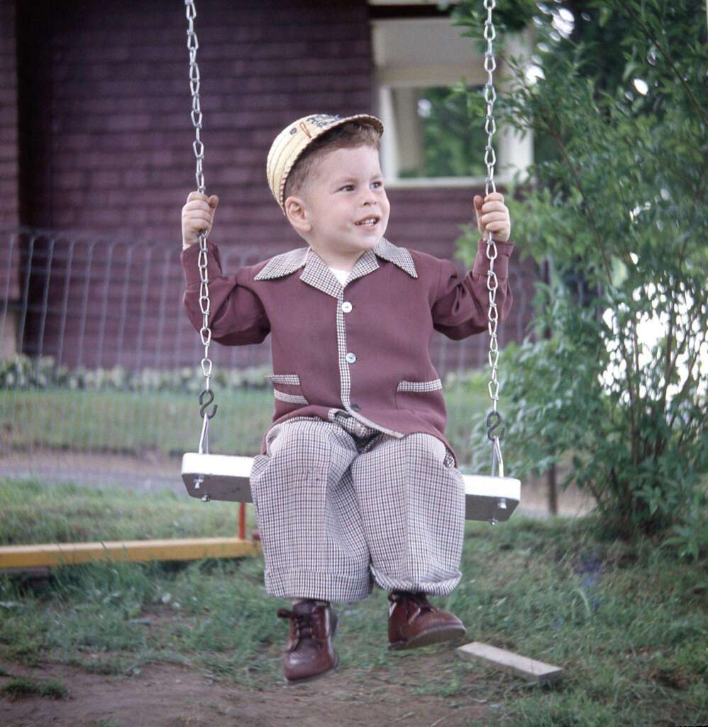 A young child wearing a brown jacket, checkered pants, and a straw hat sits on a swing outdoors. The child smiles, holding onto the chains of the swing. In the background, there is a house, greenery, and a wire fence.