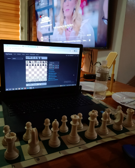 A laptop displaying an online chess game is set on a table alongside a physical chessboard with white chess pieces arranged in the starting position. A TV screen in the background shows a paused movie scene featuring a woman. A water bottle and a smartphone stand are also on the table.