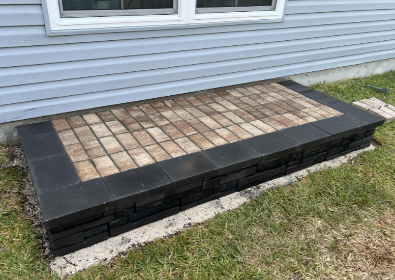 A rectangular patio step made of brown and black pavers sits against the light blue siding of a house. The step is bordered with black pavers while the inner section features a grid pattern of alternating brown and tan pavers. Grass surrounds the step.