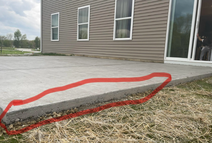 A concrete patio extends from the back of a house with beige siding and several windows. A red line is drawn around the uneven edge where the concrete meets the ground, highlighting a gap or potential issue. In the background, there is grass and some hay on the ground.