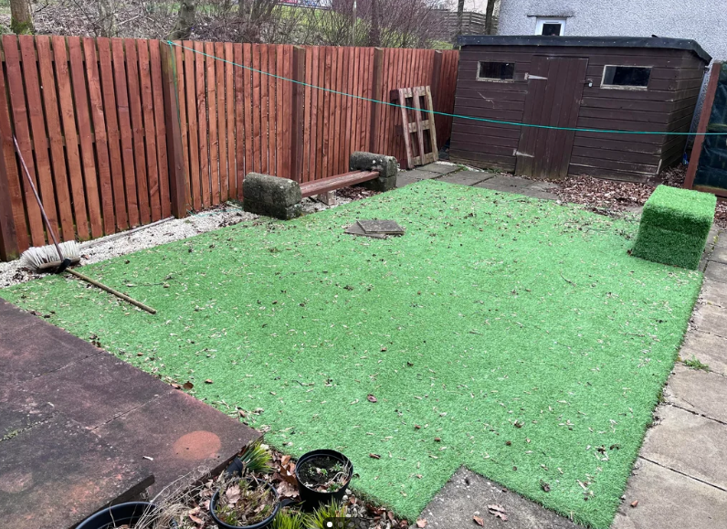 A fenced backyard features artificial grass. A brown shed is in the top right corner, and a wooden bench placed against the fence on the left. A couple of planters are in the lower left corner. Scattered logs and a broom lie on the ground.