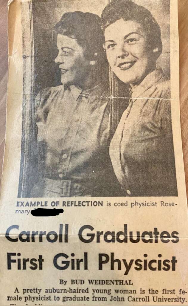 A vintage newspaper clipping features a young woman wearing a collared shirt, standing beside a mirror reflecting her image. The headline reads "Carroll Graduates First Girl Physicist." The description below mentions her graduating from John Carroll University.