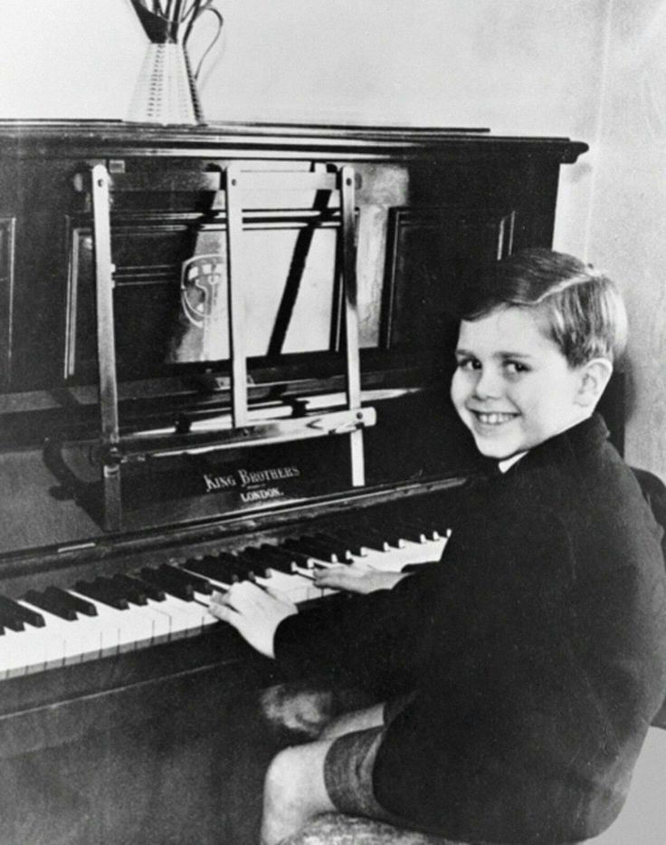 A young boy, smiling, sits at an upright piano while looking at the camera. He is dressed in a dark jacket and light-colored shorts. Behind him is a vase with flowers on top of the piano. The piano brand "Aires Brothers" is visible above the keys.