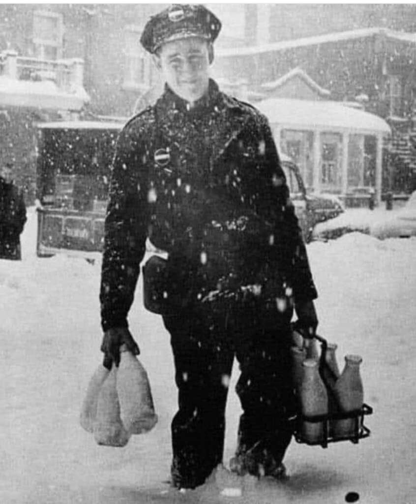 A milkman stands in the snow, smiling as he navigates a snow-covered street. He is dressed in a uniform and hat, holding a carrier with glass milk bottles in one hand and a couple of additional milk bottles in the other. Snow is actively falling around him.