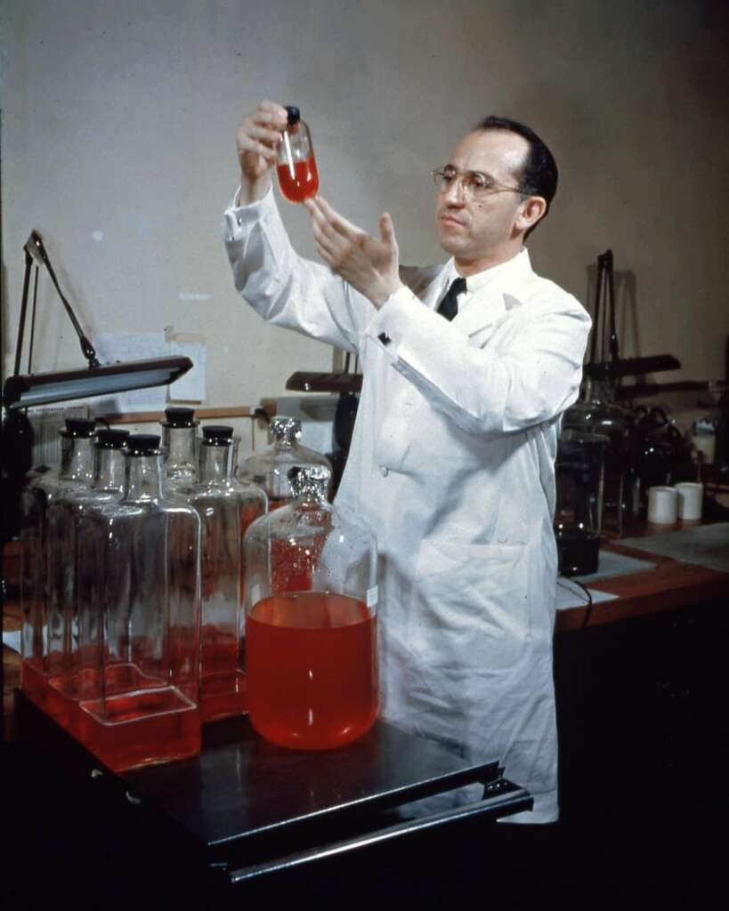 A scientist in a white lab coat holds a vial with a red liquid up to eye level, examining it intently. Several large glass bottles filled with the same red liquid are on the lab bench in front of him. The lab is equipped with various scientific apparatus.