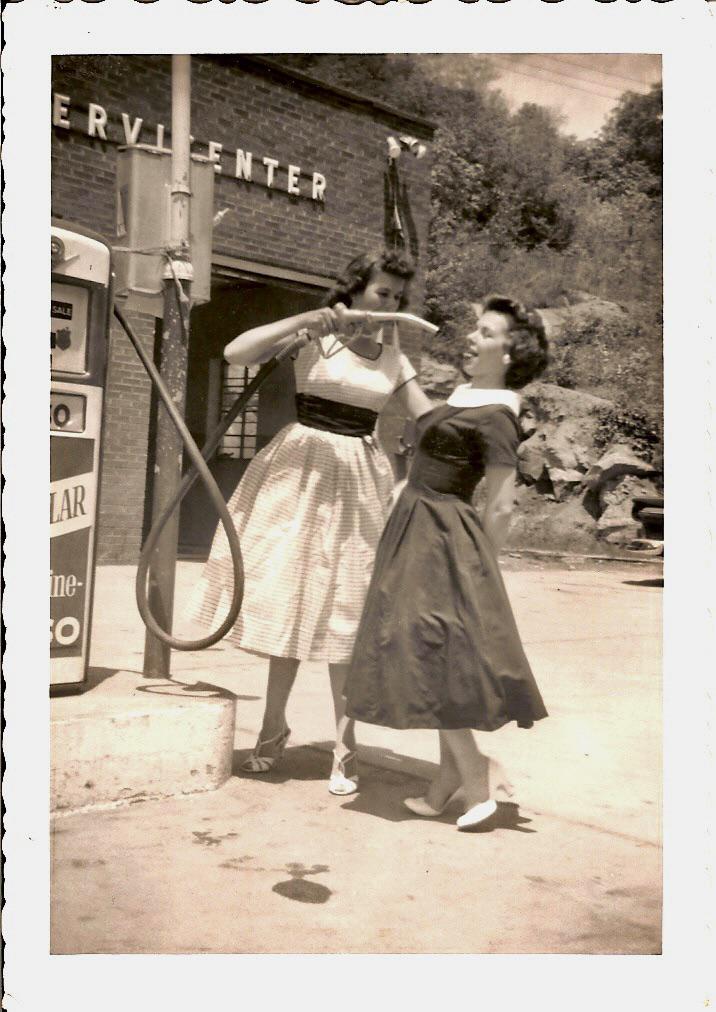 Two women are pictured at an old-fashioned gas pump. One woman playfully pretends to fuel the other with a gas nozzle, while both smile and laugh. They are dressed in vintage 1950s-style clothing, with a brick building in the background.