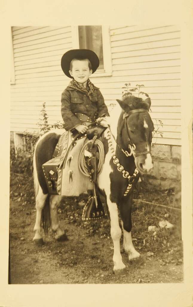 A young boy wearing a cowboy outfit, including a hat and plaid shirt, sits smiling on a small pony adorned with a saddle, bridle, and decorative tack. The background shows the side of a wooden building with horizontal siding. The image is in sepia tone.