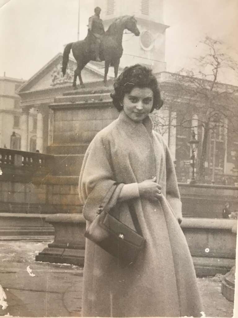 A black-and-white photo of a woman wearing a long coat and carrying a handbag, standing in front of a statue of a person on horseback. The background shows a large building with columns and a clock tower. The trees are bare, suggesting a cold season.