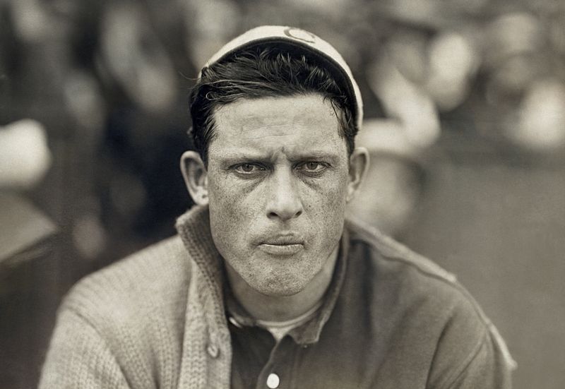 Black and white photo of a serious-looking man wearing a baseball cap and a thick cardigan over his jersey. He appears focused and determined, with the background blurred out, suggesting a sports setting.