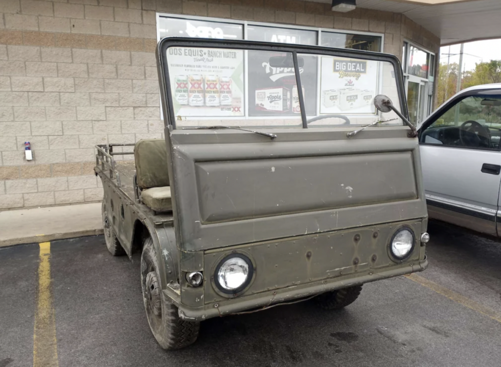 An old military-style 6-wheel vehicle parked outside a storefront. The vehicle has a boxy design with a weathered green exterior, two large round headlights, and an open-top roof. A white pickup truck is parked next to it.