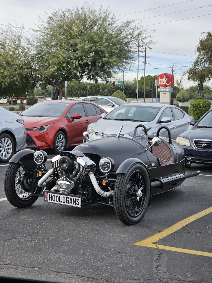 A black three-wheeled Morgan roadster with a "HOOLIGANS" license plate is parked in a parking lot. Other cars, including red and silver sedans, are visible in the background, as is a Jack in the Box sign and trees under a cloudy sky.