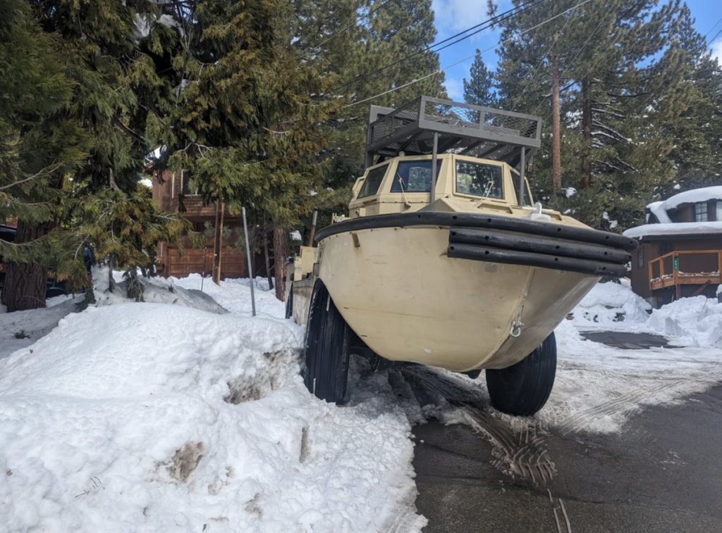 A large, beige amphibious vehicle, partially lifted on an incline of snow next to a snowy path. The background features tall evergreen trees and a wooden cabin. Tracks in the snow indicate recent movement.