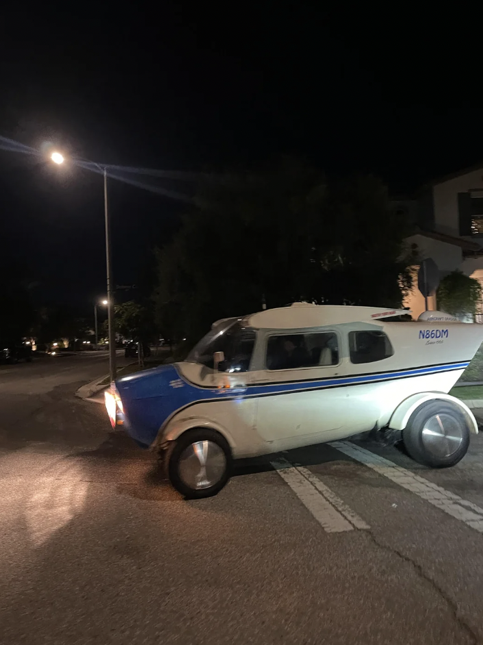 A small, unique vehicle resembling a combination of a car and an airplane drives on a street at night. The vehicle has a blue and white body with an airplane-like cockpit and tail, and is fitted with tires for road travel.