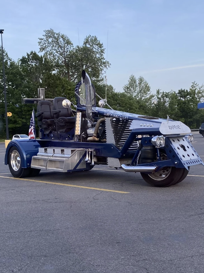 A custom-built, futuristic truck with exposed engine components, a sleek blue and silver color scheme, and "RAPTOR" written on the front bumper, parked in a parking lot with trees in the background. An American flag is attached to the rear.