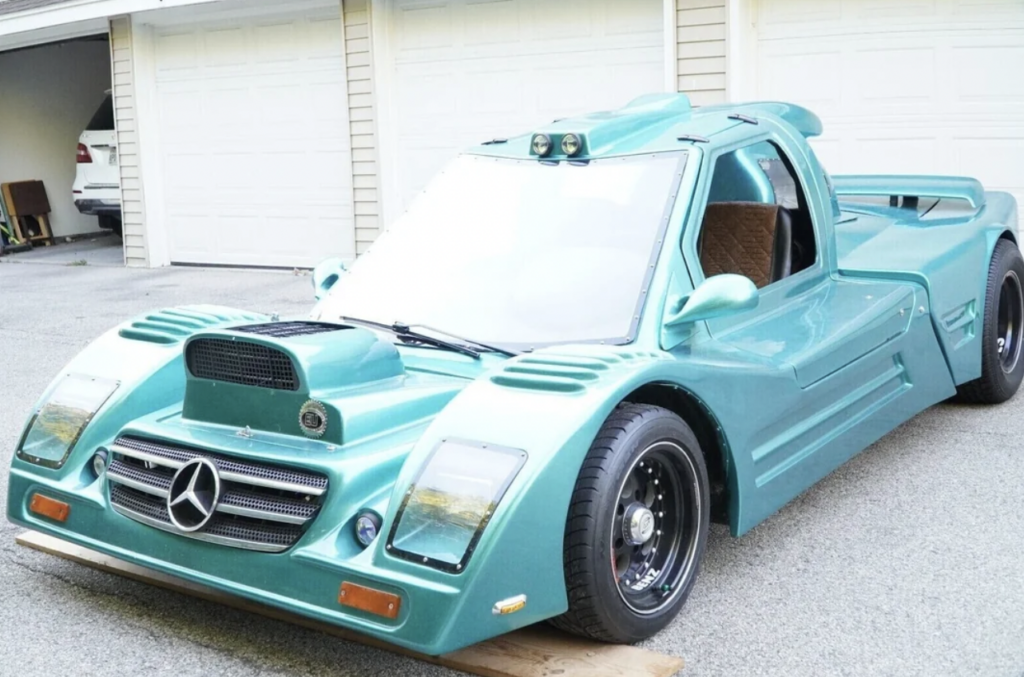 A car with a futuristic design, painted teal, and featuring a Mercedes-Benz logo on the grille, is parked in front of a beige garage. The car has distinctive features, including a large hood scoop, aerodynamic vents, and a sleek, low-profile body.