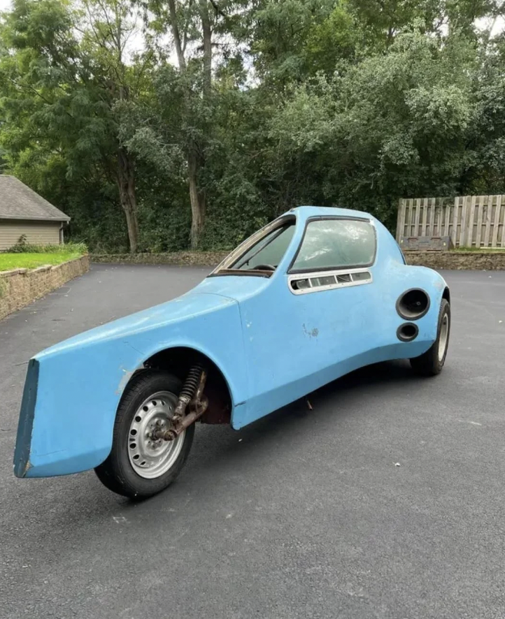 A unique, vintage blue three-wheeled vehicle with a streamlined, futuristic design is parked on a driveway. The rear wheel is missing, revealing the suspension. In the background, there are trees and a wooden fence.