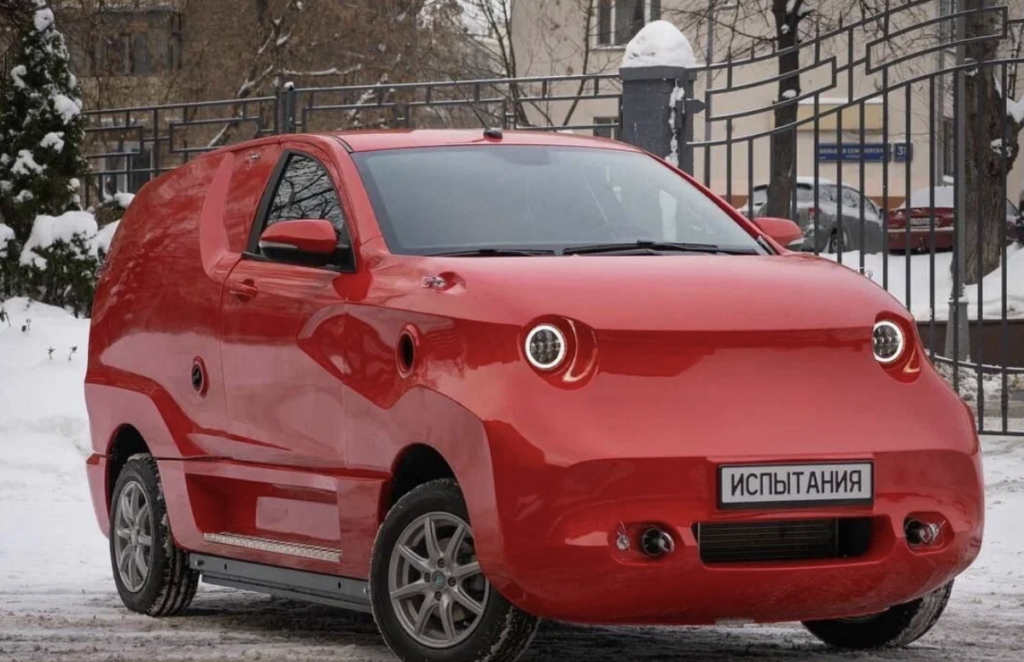 A small, red, futuristic-looking vehicle is parked on a snowy street. It has a streamlined design with round headlights, minimal windows, and a compact build. The front license plate has text in Cyrillic characters. Snow covers the ground and surrounding area.