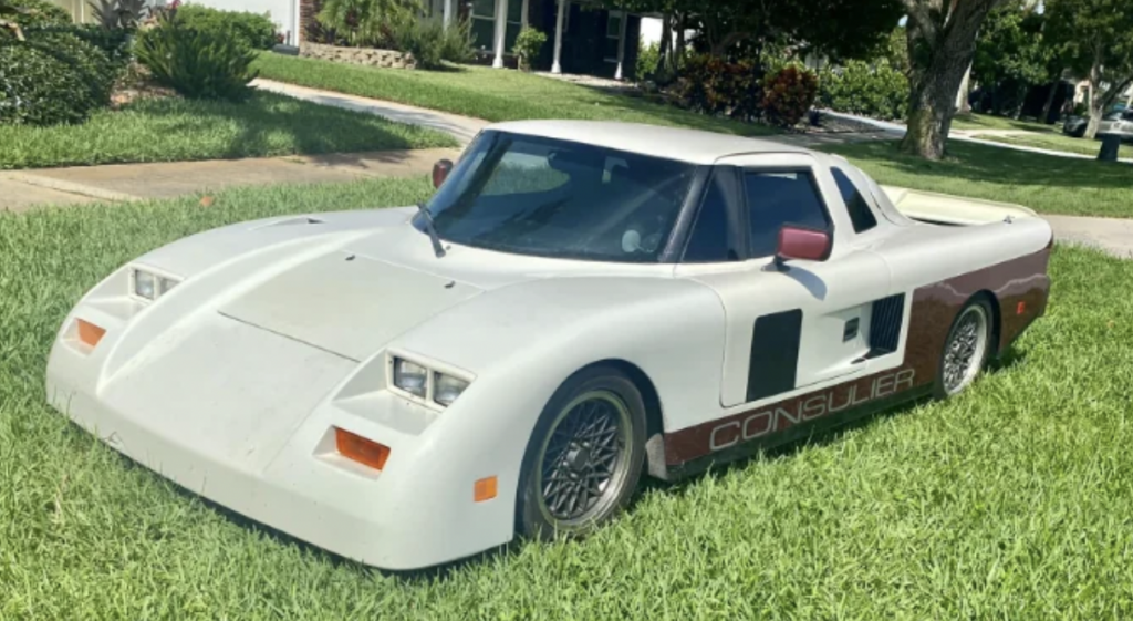 A white Consulier sports car is parked on a grassy lawn. The car has a sleek, aerodynamic design with a low profile, pop-up headlights, and a maroon accent along the bottom edge. Trees and a house are visible in the background.