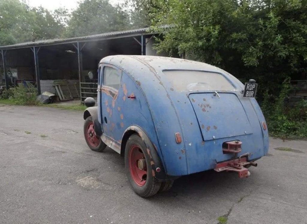 A vintage blue car with rust patches and red wheels is parked outside a shed. The vehicle has a rounded rear and a rectangular hatchback. Its license plate reads “ROP 81R.” The car is positioned on a mostly empty paved area surrounded by trees and other structures.