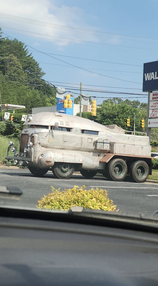 A large, custom-built vehicle resembling a futuristic armored transporter is stopped at a traffic light. The vehicle has a rusty, weathered exterior and six wheels. It is seen from a car's interior, with traffic lights and greenery in the background.