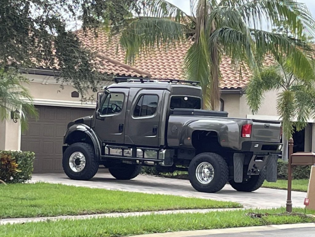 A large, dark gray custom pickup truck with an extended cab is parked in the driveway of a suburban home with a red-tiled roof. Palm trees and greenery surround the area, and there's a mailbox visible in the bottom right corner of the image.