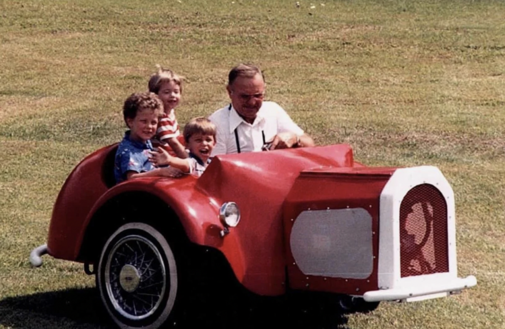 An elderly man is driving a small, red, vintage-style car with three young boys. They are all smiling and appear to be enjoying a sunny day outside on a grassy field. The car has retro design elements and the overall scene is lighthearted and playful.