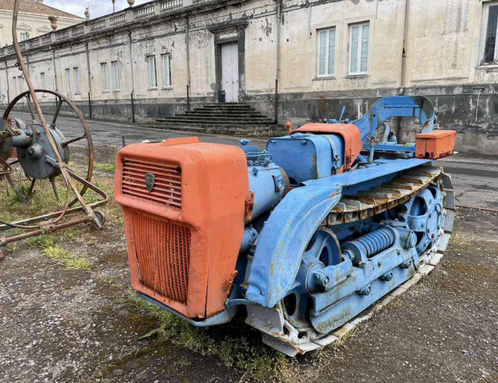 A weathered, blue and orange tracked tractor is parked on a gravel surface in front of a large, old, beige building with several windows and a central door. The tractor shows signs of rust and wear, indicating it has not been used for some time.