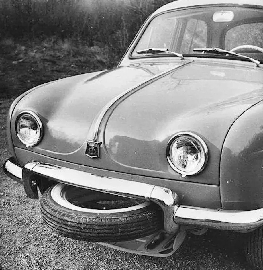 A vintage car with a spare tire mounted on its front bumper. The car has a rounded hood with two round headlights and a sleek, simplistic design, reflecting automotive styles of the mid-20th century. The background shows a blurred natural setting.