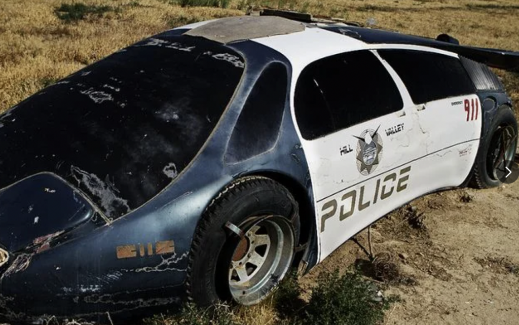 A weathered, futuristic-looking police car with "Police" and "911" on its side is abandoned in a dry, grassy area. The car, which has a white and black paint job, is missing its front bumper and has cracked windows, appearing neglected and damaged.