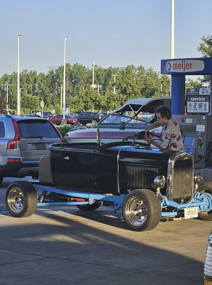 A person standing near a classic black hot rod car with blue frame at a gas station. Trees and parked cars are visible in the background, along with a Meijer sign and gas pumps.