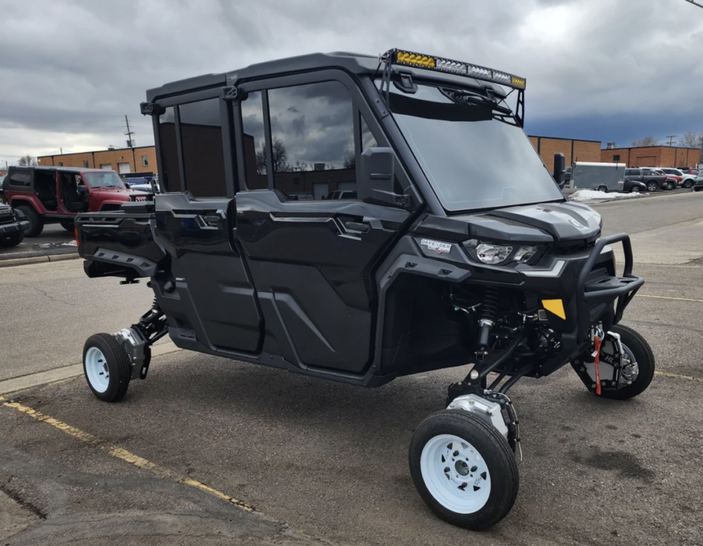 A black all-terrain vehicle (ATV) with a fully enclosed cabin and raised suspension is parked in a lot. The ATV features robust off-road tires, white rims, a winch on the front bumper, and an LED light bar on the roof. Buildings and other vehicles are in the background.