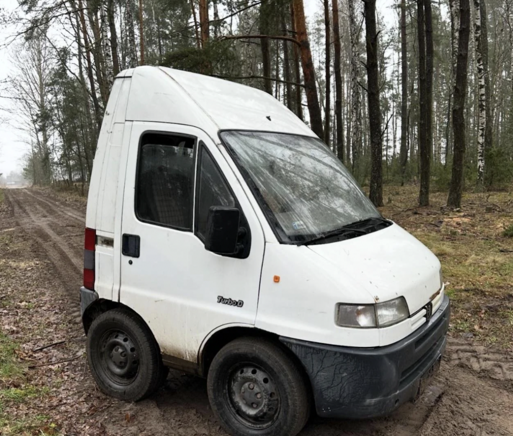 A white van with a compact body and three wheels is parked on a dirt path in a forested area. The vehicle has a tall, narrow design. Trees with sparse foliage surround the path, and the ground is wet and muddy.