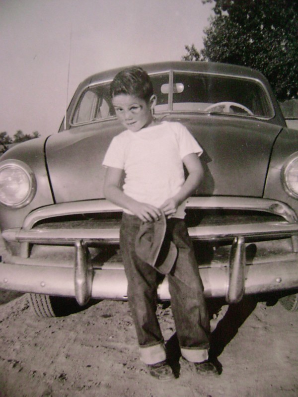 A young boy wearing a white t-shirt, dark jeans, and sneakers stands in front of a vintage car, holding a hat in his right hand. The photo is in black and white, and the boy is smiling slightly while looking off to the side. Trees are visible in the background.