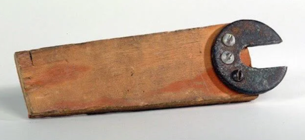 A rectangular piece of wood with a rusted, metal crescent-shaped clamp attached to one end. The clamp is fastened to the wood with three visible screws, giving the appearance of a makeshift or vintage tool. The background is plain white.