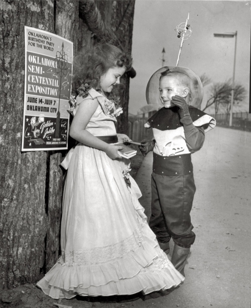 A black-and-white photo shows a young girl in a dress and a boy in a space-themed costume. They stand next to a tree with a vintage poster for the "Oklahoma Semi-Centennial Exposition" held in Oklahoma City from June 14 to July 7. The girl is handing something to the boy.