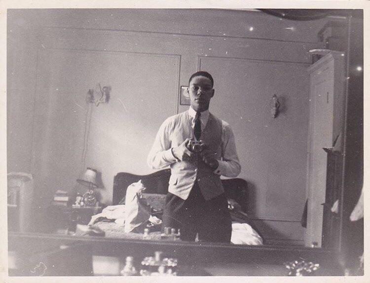 A black-and-white photo shows a man taking a selfie in a mirror. He is dressed in a shirt, tie, and vest. The room in the background is slightly cluttered, with a bed, bedside table with a lamp, and various items scattered around.