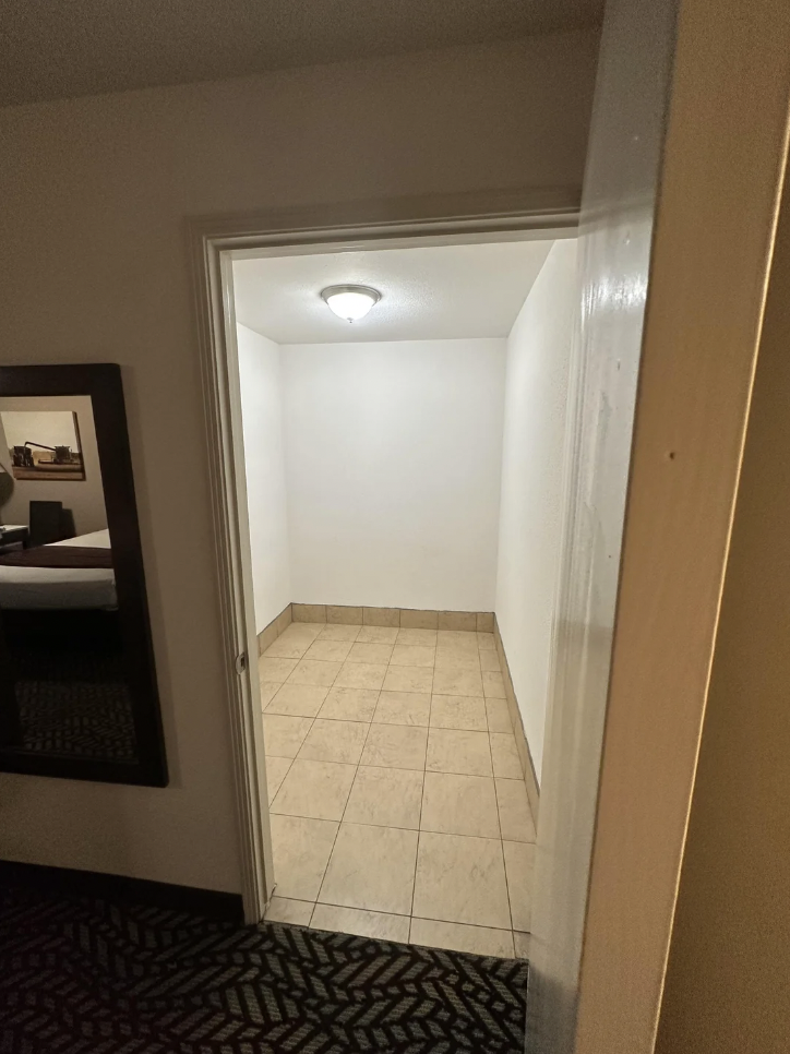 A small, empty room with white walls and a tiled floor, viewed through an open doorway. The room is illuminated by a ceiling light. A partial view of a mirror on a wall in the adjacent room is visible on the left side of the image.