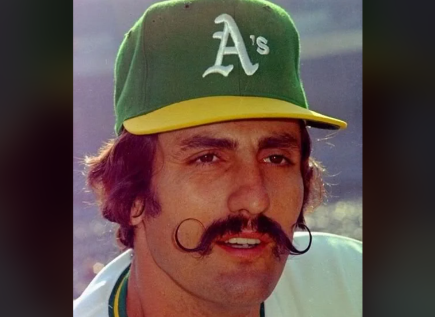 A person with a handlebar mustache and sideburns is wearing a green baseball cap with a white "A's" logo on the front and a yellow brim. They are also dressed in a white and green baseball uniform. The background is blurred.