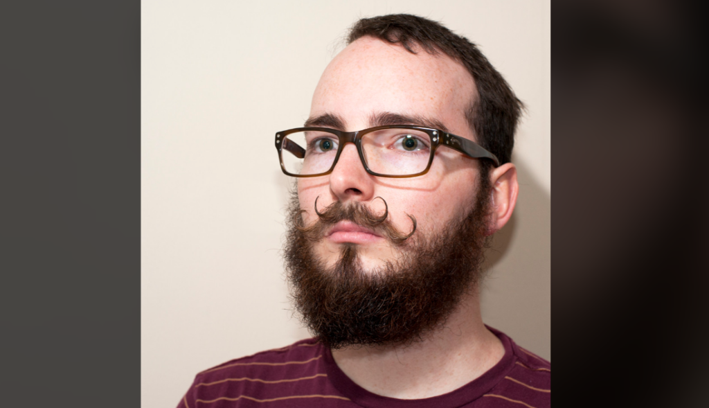 A man with short dark hair and glasses is looking slightly to the left. He has a full beard and a styled mustache with curled ends. He's wearing a maroon striped shirt and is standing against a neutral background.