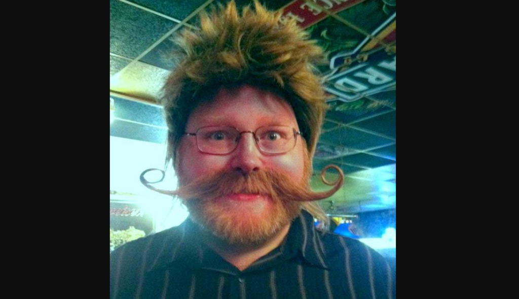 A man with reddish hair and a full beard wears a spiky wig and has a handlebar mustache curled at the ends. He is wearing glasses and a striped shirt. The background shows a ceiling with posters and signs, suggesting a lively indoor setting.