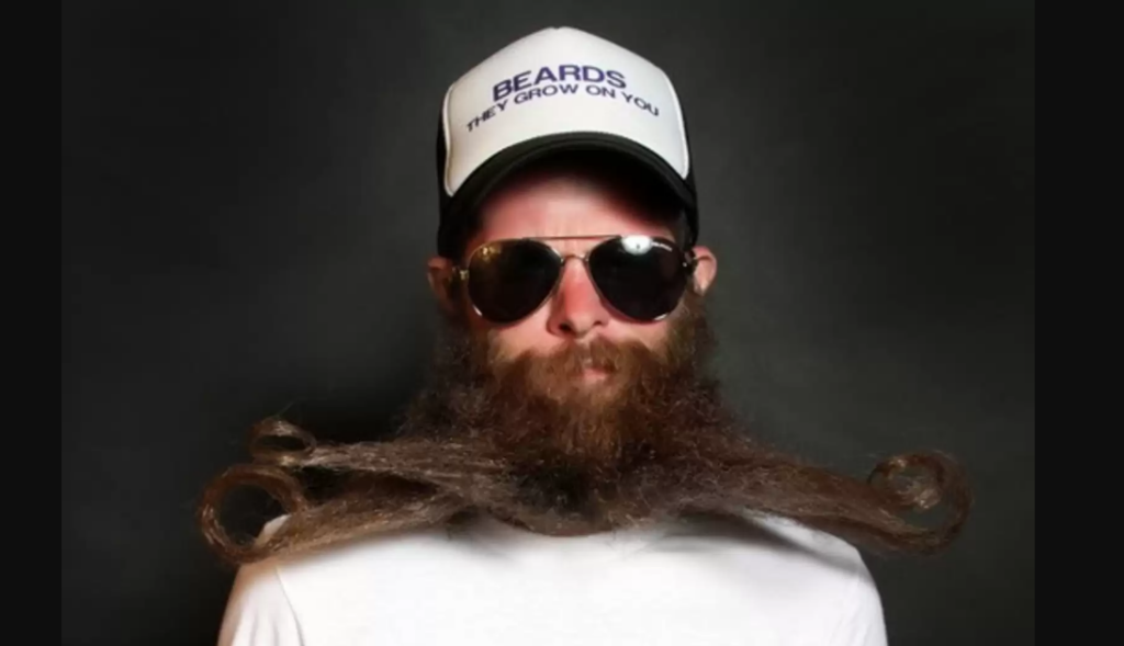 A person with a long, intricately styled beard that curls at the tips is wearing a white baseball cap that reads "BEARDS - THEY GROW ON YOU" and aviator sunglasses. The person is dressed in a simple white t-shirt with a black background.