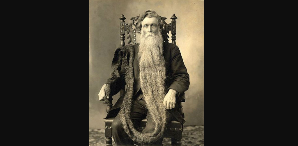 A black and white photo of an elderly man sitting in an ornate wooden chair. He has an exceptionally long beard that extends down to his lap and beyond, draping over the side of the chair. The man is dressed in a dark suit and has a serious expression on his face.