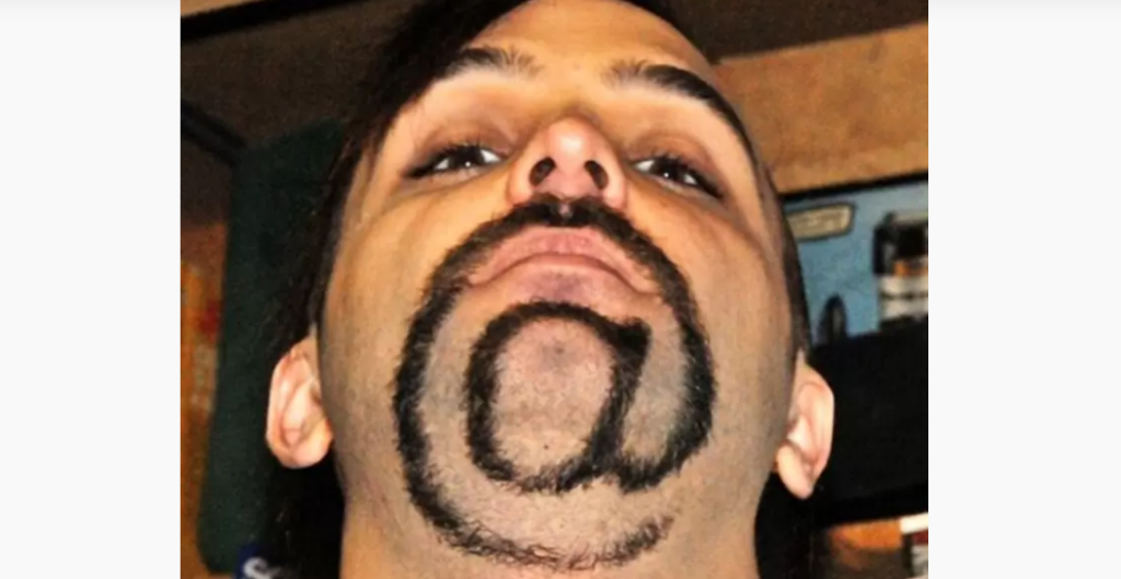 Portrait view of a person looking up, revealing a unique beard style. The facial hair is groomed to form the shape of the "@" symbol, encompassing the chin and extending up towards the cheeks. The background is blurred and indistinct.