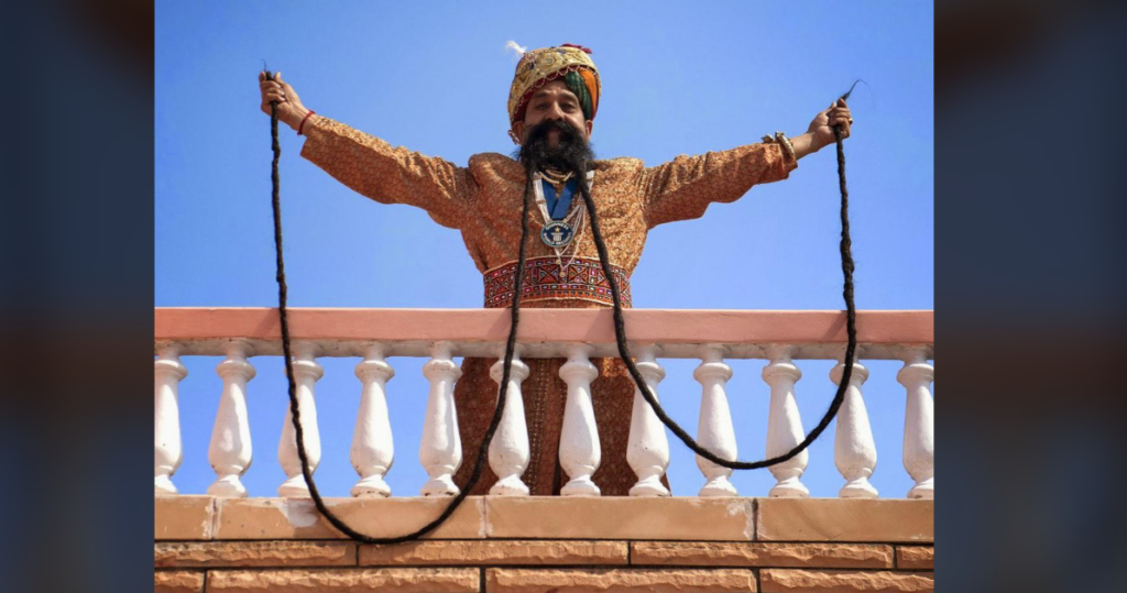 A person dressed in ornate traditional attire with a turban stands on a balcony with outstretched arms, holding long strands of hair, creating a loop. The clear blue sky serves as the background.