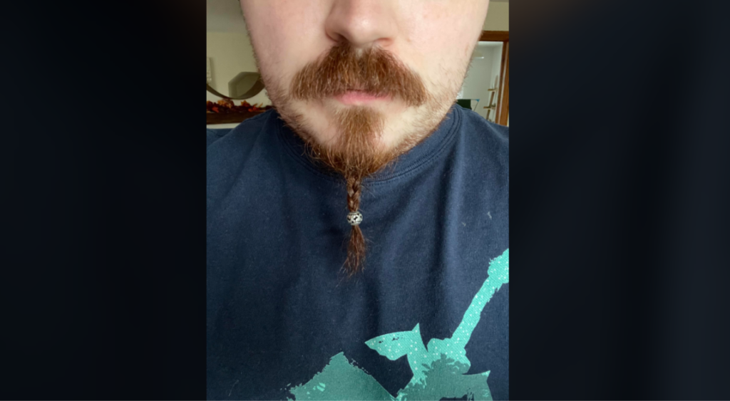 Close-up of a person with a brown mustache and a small, braided beard with a silver bead. The person is wearing a navy blue shirt with a turquoise graphic on it. Background details are out of focus.